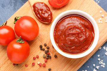 Board with bowl of tasty tomato paste, spoon, peppercorns and salt on blue grunge background