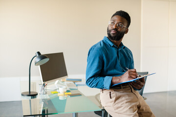 Portrait of thoughtful black man standing in office near workplace, holding clipboard and looking aside at free space