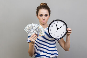 Woman holding wall clock and fun of dollar banknotes, looking at camera.