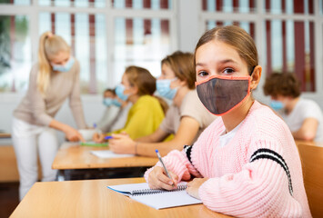 Fototapeta premium School girl in face mask sitting at desk in classroom on background with classmates and teacher