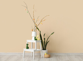 Vase with tree branches, Easter eggs and toy rabbits on stepladder near beige wall in room