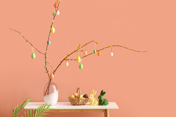 Vase with tree branches, Easter eggs and toy rabbits on table near color wall