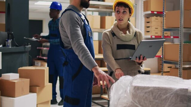 Caucasian people analyzing list of goods on tablet, working in warehouse with retail merchandise and boxes. Supervisor giving tasks to worker, organizing products on shelves and racks.