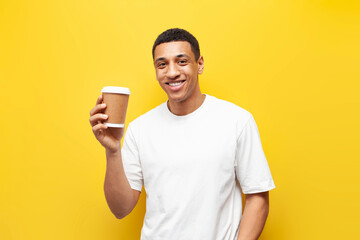 young african american guy in white t-shirt holds and recommends coffee in paper cup on yellow background