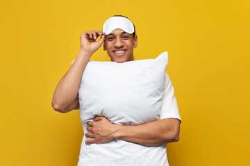 young african american guy in pajamas and sleep mask hugs soft pillow on yellow isolated background