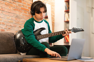 Asian Boy Playing Guitar Wearing Earphones Using Laptop At Home