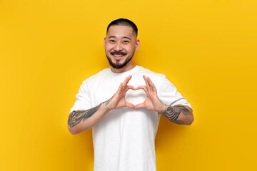 adult tattooed asian man in white t-shirt shows heart with his hands on yellow isolated background...