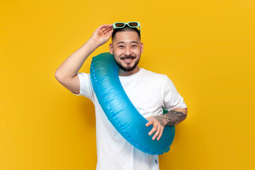 tattooed male asian tourist in white t-shirt and sunglasses holds inflatable swim ring and goes on...