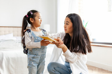 asian mom gives book to daughter and kisses her at home, little girl going to study with mom, back to school