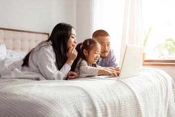 asian family with little daughter lie on bed at home and use laptop, korean dad mom and daughter look at computer screen