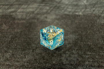 Blue twenty-sided resin die with gold inclusions on a wooden table, all numbers inked white except golden 20