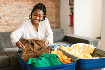 Positive black woman packing suitcase for vacation, sitting near open suitcase and preparing for...