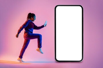 Young african american woman exercising, running with cellphone next to giant smartphone with blank screen, mockup