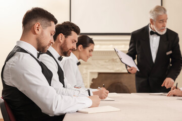 Senior man in formal suit teaching trainees indoors. Professional butler courses