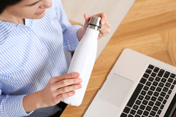 Young woman holding thermo bottle at workplace indoors, above view