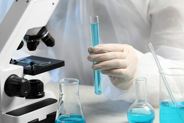 Scientist with test tube of light blue liquid near microscope in laboratory, closeup