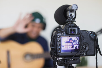 Video production on professional camera recording young Asian man playing guitar and greeting fans at home studio