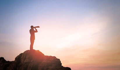 Businessman with binoculars on peak of Mountain. Looking Far Away against sunset Sky background. Concept of business man's Vision, Perspective and Future Planning. 