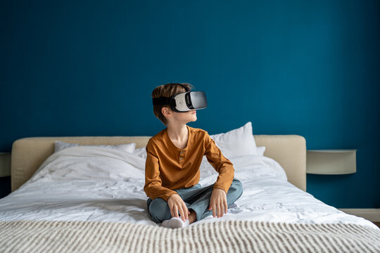 Kid interact using virtual reality headset to interact with the imaginary world. Caucasian boy child sitting on bed at home wearing VR glasses playing video games. Immersive learning and children