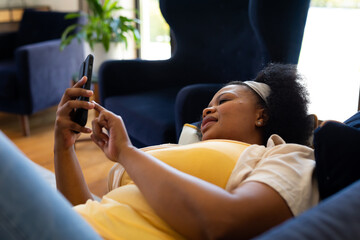 Happy plus size african american woman lying on sofa and using smartphone