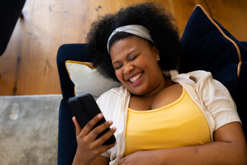 Happy plus size african american woman lying on sofa using smartphone and laughing