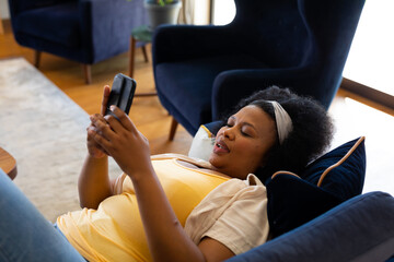 Happy plus size african american woman lying on sofa and using smartphone