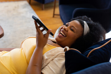 Happy plus size african american woman lying on sofa, talking on smartphone and laughing
