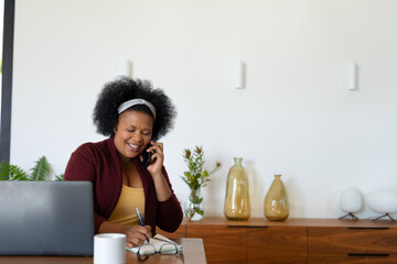 Happy plus size african american woman at table with laptop, taking notes and talking on smartphone
