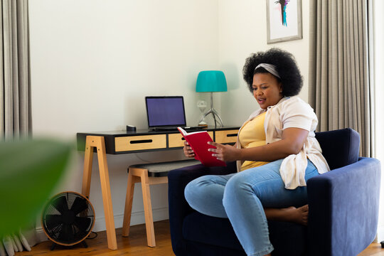 Happy plus size african american woman sitting in armchair at home, reading book