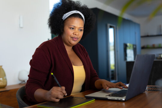 Happy plus size african american woman sitting at table, using laptop and drawing tablet - Powered by Adobe