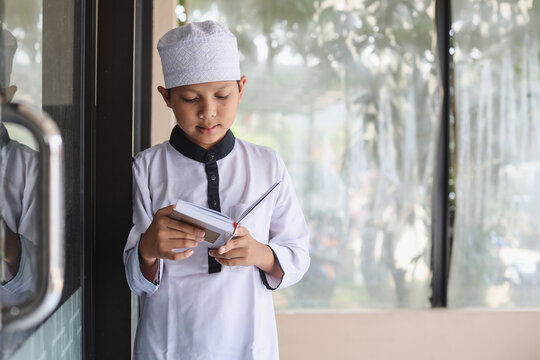 Muslim boy in prayer cap and arabic clothes reading holy quran book praying to Allah, ramadan kareem concept kid spiritual peaceful moment.