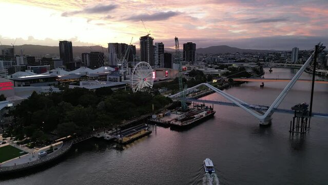 Drone shot of South Bank, shot over Brisbane River, camera orbits The Wheel of Brisbane. Citycat Boats approaching South Bank pontoon. New Neville Bonner Bridge in construction in background, Sunset.