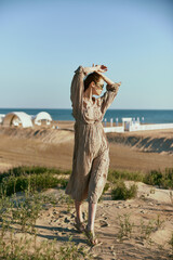 a slender, sophisticated woman in a beige dress stands on the sand in windy weather with her hands raised to her head