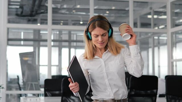 Cheerful funny businesswoman dances with green wireless headphones on, holding a cup of coffee and a folder of documents in office. Female playful energy demonstrate the importance of taking breaks.