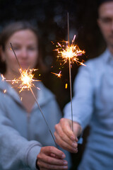 Female and male hands holding sparkling fireworks at twilight against trees in the evening. Couple celebrating holiday - 586045411