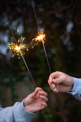 Female and male hands holding sparkling fireworks at twilight against trees in the evening. Couple celebrating holiday - 586045413