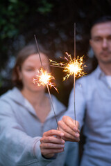 Female and male hands holding sparkling fireworks at twilight against trees in the evening. Couple celebrating holiday - 586045491