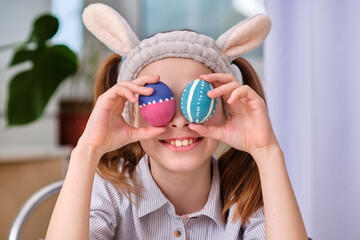 A positive playful girl in bunny ears holds colorful eggs in her hands in the home kitchen. Happy Easter. Easter background