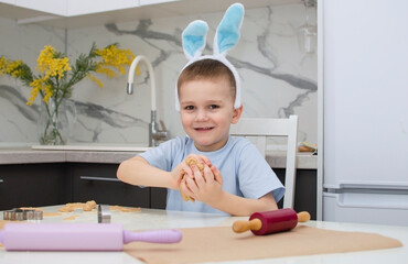 An emotional happy little child with bunny ears smiles and bakes cookies for Easter at home in kitchen. Happy Easter. Home bakery