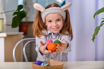 A positive playful girl in bunny ears holds colorful eggs in her hands in the home kitchen. Happy Easter. Easter background