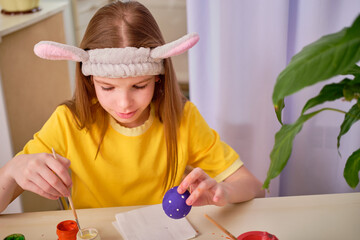 A child paints Easter eggs with paints. Portrait of caucasian girl, child 9-10 years old decorating easter eggs in the domestic kitchen. The concept of Easter