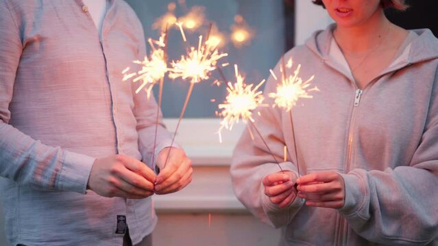 Female and male hands holding sparkling fireworks at twilight against trees in the evening. Couple celebrating holiday