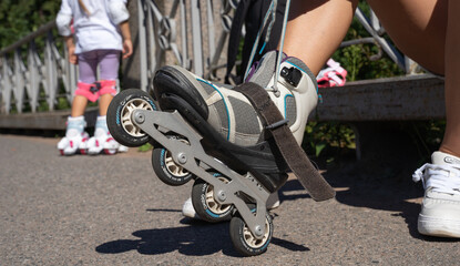 Plakat Close-up Mom putting on inline skates rollers preparing with child in public park in summer. Family leisure outdoor sport activity game.