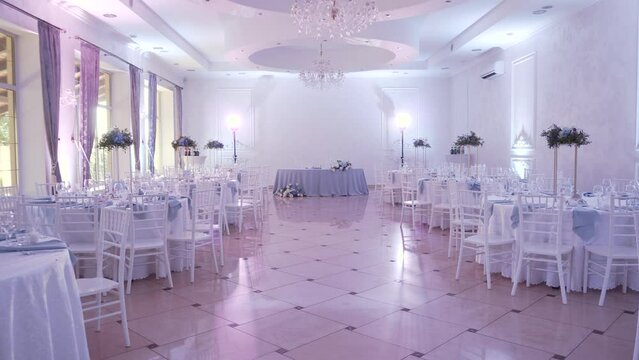 Beautiful rich large banquet hall decorated with flowers with round tables, white chairs with professional blue and white spotlights. Tiled floors, crystal chandeliers and large windows on wall.