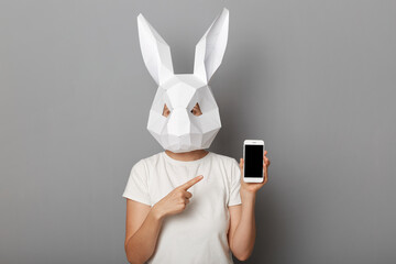 Portrait of unrecognizable woman wearing white t shirt and paper rabbit mask standing isolated over gray background, showing smart phone with empty screen, pointing at copy space.