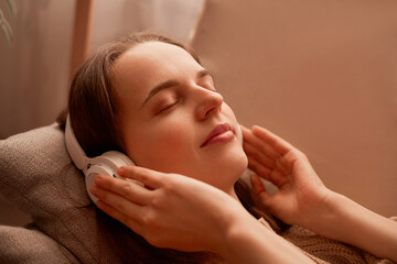 Fototapeta na wymiar Side view portrait of relaxed young adult woman listening music while resting on couch at home, keeps eyes closed, touching her headphones, enjoying moment.
