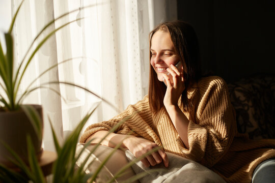 Image of young beautiful woman talking on smartphone with happy expression at home near window in sunlight, looking away with smile, hearing pleasant news or has romantic conversation.
