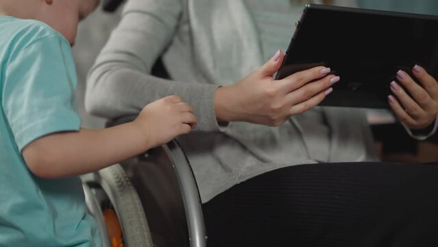 Curious toddler in blue t-shirt watches mommy checking mail on modern tablet. Woman with paralysis sits in wheelchair using electronic device closeup