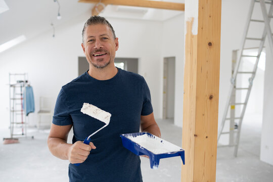 Middle-aged man with grey hair painting wooden beams in attic apartment, construction site with white paint