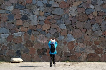 Woman in blue jacket is standing in front of colorful stone wall and taking photo